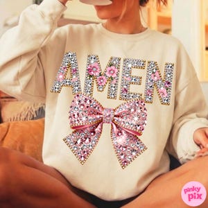 May include: A cream-coloured sweatshirt featuring the word "AMEN" in jewelled letters with pink floral details. A large, sparkling pink bow is positioned below the text. The sweatshirt is a relaxed, comfortable style.