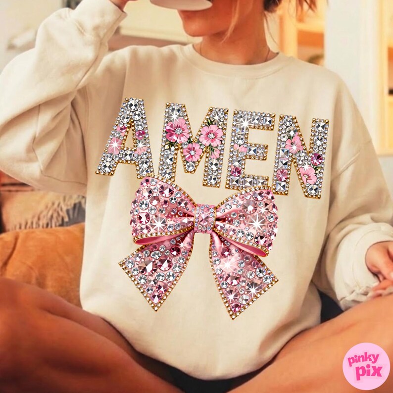 May include: A cream-coloured sweatshirt featuring the word "AMEN" in jewelled letters with pink floral details. A large, sparkling pink bow is positioned below the text. The sweatshirt is a relaxed, comfortable style.