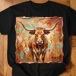 May include: Black t-shirt featuring a colorful, textured painting of a longhorn steer. The artwork uses shades of brown, orange, and teal, with long curved horns and a detailed face. The shirt is a classic crew neck.