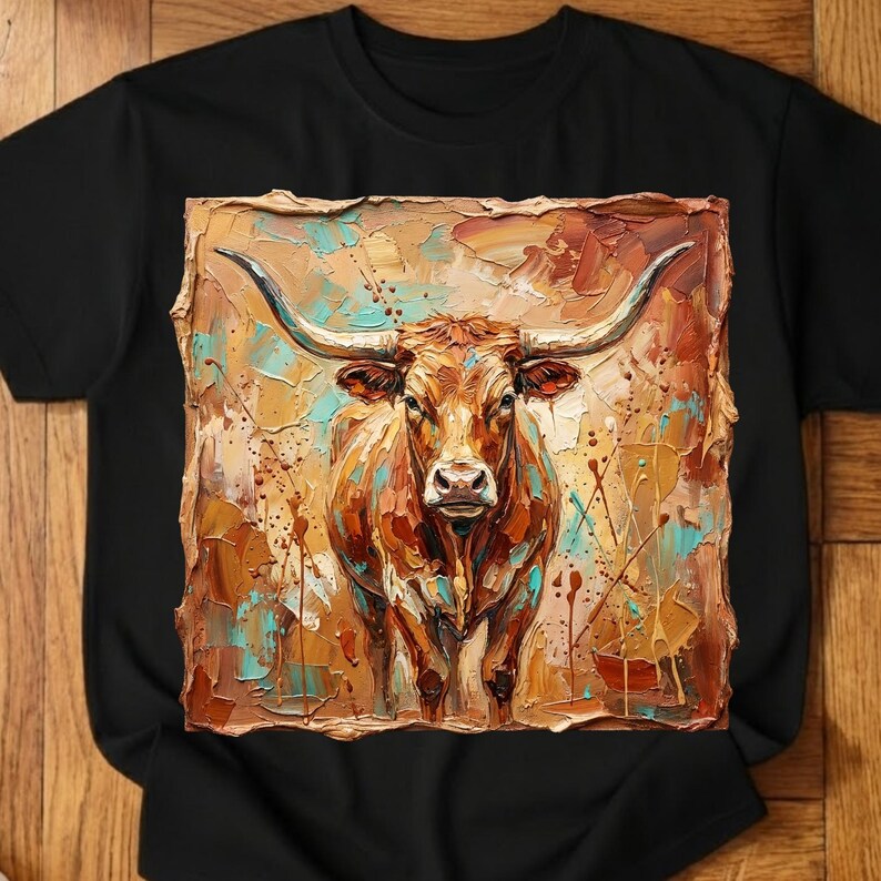 May include: Black t-shirt featuring a colorful, textured painting of a longhorn steer. The artwork uses shades of brown, orange, and teal, with long curved horns and a detailed face. The shirt is a classic crew neck.