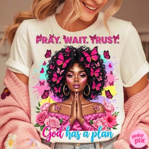 May include: White t-shirt featuring a graphic of a woman with butterflies and the text "PRAY. WAIT. TRUST." at the top. The words "God has a plan" are at the bottom. The background includes pink and yellow paint splatters.