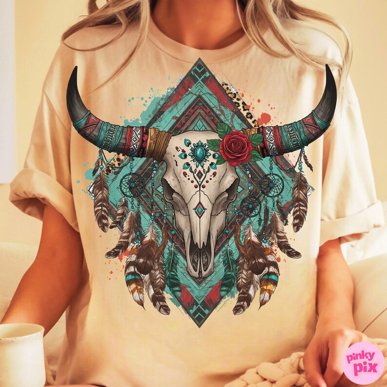 May include: Cream-coloured t-shirt with a detailed graphic of a bull skull with decorative horns. The design includes turquoise, red, and brown accents, feathers, and a red rose. The shirt has a relaxed fit.