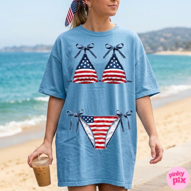 May include: Pale blue t-shirt with a bikini design in the style of the American flag. The bikini top and bottom are red and white striped with blue star details. The shirt has short sleeves and a loose fit, ideal for warm weather.