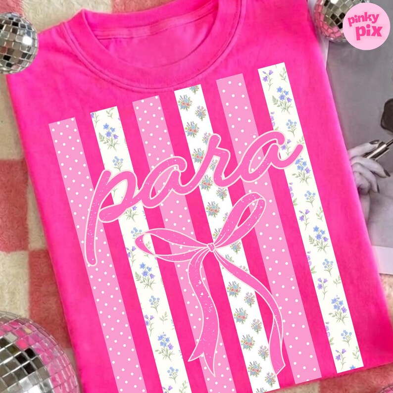 May include: A bright pink t-shirt with a graphic design. The design features vertical stripes in pink and white, with polka dots and floral patterns. The word "para" is written in a cursive font, and a bow is at the bottom.