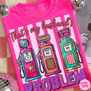 May include: A bright pink t-shirt with the phrase "That's my husband's PROBLEM" and illustrations of three vintage gas pumps. The gas pumps are in shades of pink, orange, and teal.