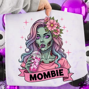May include: A white print featuring a zombie woman with pink and blue hair, wearing a pink shirt and flower crown, drinking from a pink cup. The word "MOMBIE" is on a pink banner below the image. Pink stars are scattered around the image.