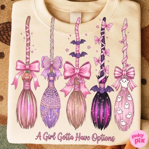 May include: Cream-colored sweatshirt featuring five decorative witch brooms in shades of pink, purple, and brown. Each broom has unique embellishments, including bows, bats, spiders, and ghosts. The text "A Girl Gotta Have Options" is printed at the bottom.