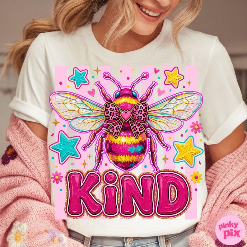 May include: White t-shirt featuring a colorful graphic of a bee with a bow and the word "KIND" in pink. The bee has pink, yellow, and black stripes. The design includes stars and flowers. The shirt is part of a clothing collection.