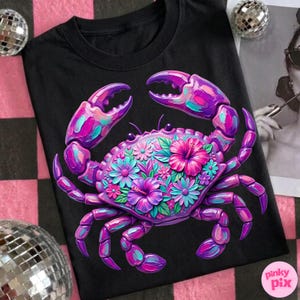 May include: Black t-shirt featuring a vibrant, stylized crab design. The crab is rendered in shades of purple, pink, and teal, adorned with floral accents. The design includes a variety of flowers, including hibiscus. The shirt has a crew neck.