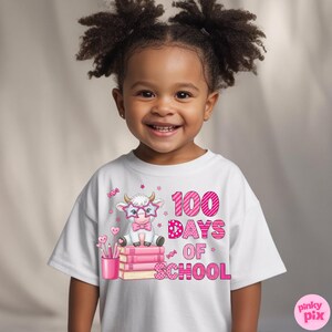 Pu&ograve; includere: Una maglietta bianca con una mucca dei cartoni animati con occhiali e papillon, seduta su libri rosa. Il testo "100 Days of School" &egrave; stampato in rosa. La ragazza sorride.