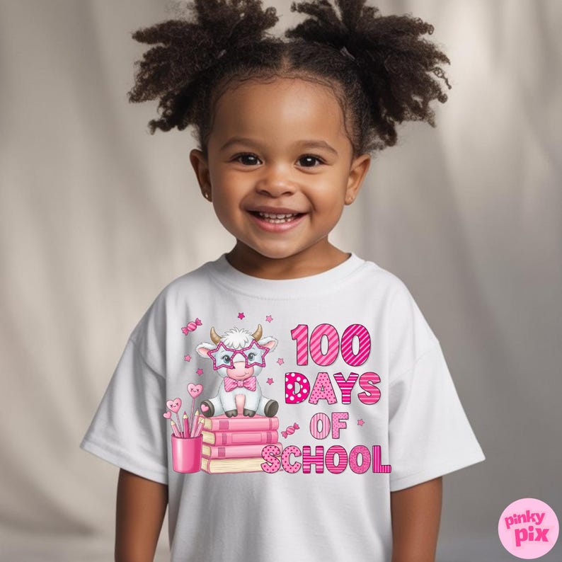 Pu&ograve; includere: Una maglietta bianca con una mucca dei cartoni animati con occhiali e papillon, seduta su libri rosa. Il testo "100 Days of School" &egrave; stampato in rosa. La ragazza sorride.