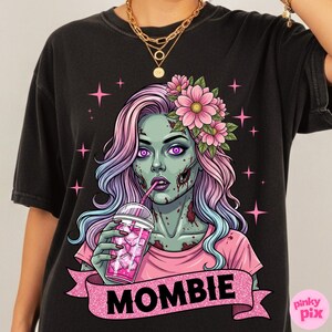 May include: Black t-shirt featuring a zombie illustration with pink and purple hair, flowers, and a drink. The zombie has green skin and the word "MOMBIE" on a pink banner.