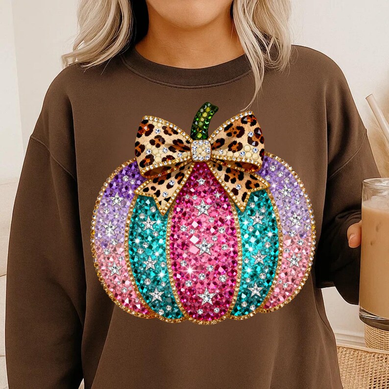 Op de afbeelding: Een bruine sweater met ronde hals en een grote afbeelding van een pompoen versierd met roze, paarse en turquoise strass-steentjes en sterren. De pompoen heeft een groene steel en een strik met luipaardprint en strass-steentjes.