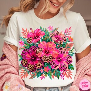 May include: White t-shirt featuring a vibrant floral design. The design showcases large pink hibiscus flowers with leopard print centers, surrounded by green leaves and smaller pink flowers. The shirt has rolled-up sleeves.
