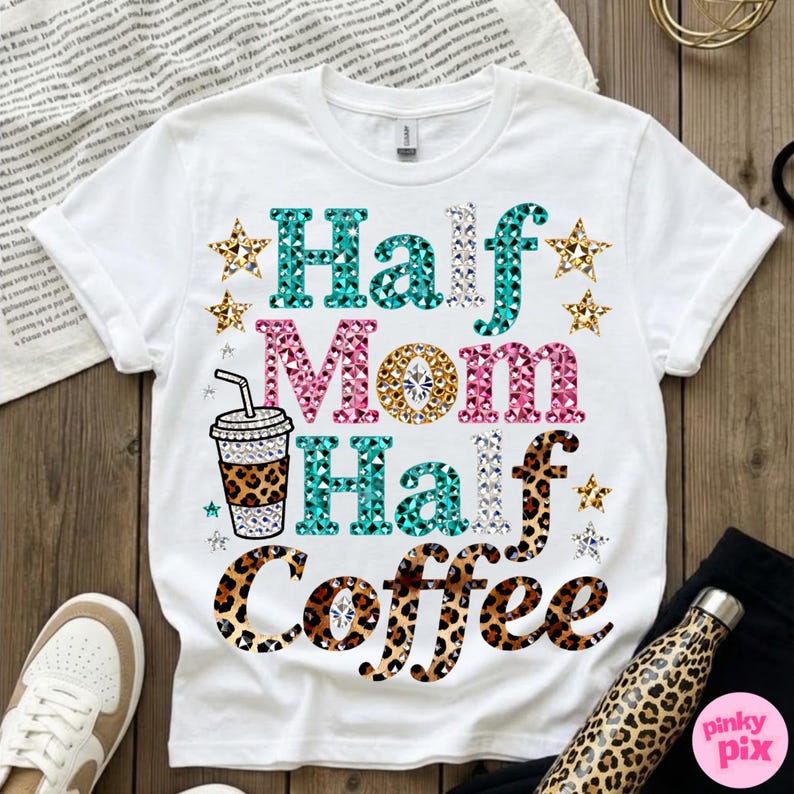 Może przedstawiać: Biała koszulka z napisem "Half Mom Half Coffee" w kolorze turkusowym, r&oacute;żowym i w panterkę. Projekt zawiera grafikę filiżanki kawy i akcenty gwiazd. Koszulka jest prezentowana na drewnianej powierzchni.