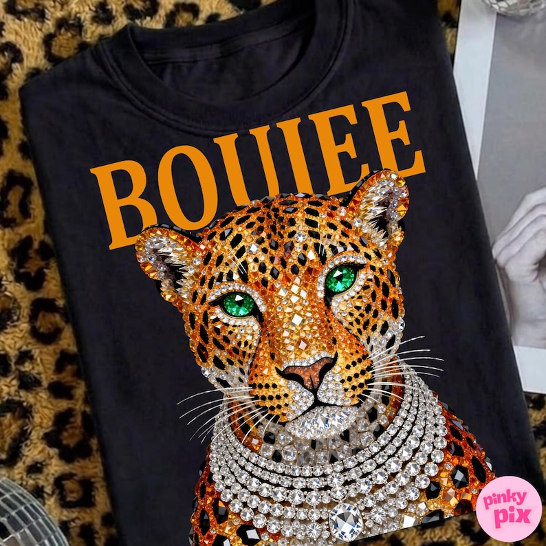May include: Black crew neck t-shirt featuring a jeweled leopard design with emerald green eyes and a diamond necklace. The word "BOUIEE" is printed in orange across the top. The leopard has a gold and brown spotted pattern.