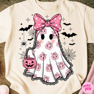 May include: Cream-coloured t-shirt with a cute ghost design. The ghost wears a pink bow with white stars and carries a pink pumpkin bucket. Black bats, spiderwebs, and stars decorate the shirt, perfect for Halloween.