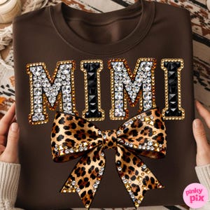 May include: A brown sweatshirt featuring the word "MIMI" in a jeweled design with a leopard print bow. The letters are outlined in gold and filled with clear and black rhinestones. The bow is adorned with rhinestones.