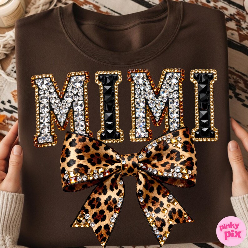 May include: A brown sweatshirt featuring the word "MIMI" in a jeweled design with a leopard print bow. The letters are outlined in gold and filled with clear and black rhinestones. The bow is adorned with rhinestones.