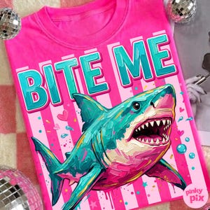 May include: A hot pink t-shirt with the words "BITE ME" in teal letters. A teal and pink shark graphic is centered below the text. The shirt has a crew neck and short sleeves. The design includes hearts and bubbles.