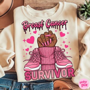 May include: Cream-colored sweatshirt with the words "Breast Cancer Survivor" in pink. The graphic features a raised fist with long pink nails, pink hearts, and pink sneakers. The design includes pink ribbons.