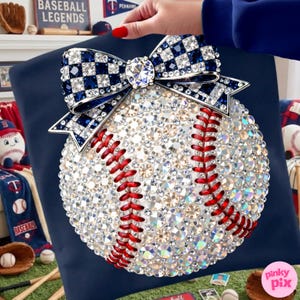 May include: A baseball-themed decorative item. The baseball is covered in sparkling crystals with red stitching. A large bow with blue and white checkered crystals sits atop the baseball. The item is on a navy blue surface.