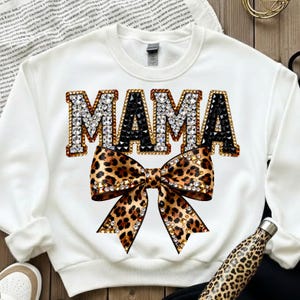 Pu&ograve; includere: Felpa bianca con la scritta "MAMA" in lettere gioiello e un fiocco leopardato. Le lettere sono bordate in oro e nero, con gioielli trasparenti e neri. Il fiocco &egrave; anch'esso ornato di gioielli.