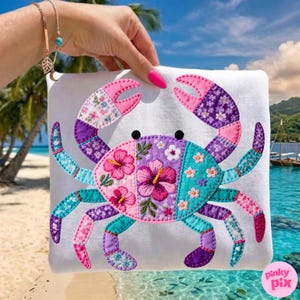 Könnte beinhalten: Ein weißes quadratisches Kissen mit einem farbenfrohen Krabbenmuster. Die Krabbe besteht aus Patchwork-Stoff in Rosa, Lila und Türkis, mit gestickten floralen Details. Die Scheren und Beine der Krabbe sind mit verschiedenen gemusterten Stoffen segmentiert.