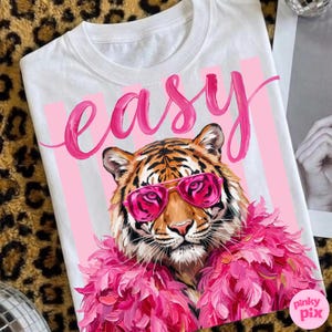 May include: White t-shirt featuring a tiger wearing pink sunglasses and a pink feather boa. The word "easy" is written in pink script above the tiger. Pink vertical stripes are behind the tiger.