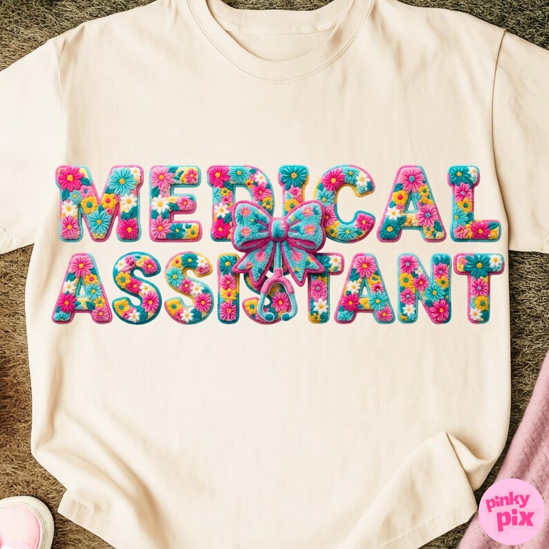 May include: Cream-colored t-shirt with the words "MEDICAL ASSISTANT" in colorful floral letters. A large bow and stethoscope graphic are centered on the shirt. The shirt is made of a soft material.