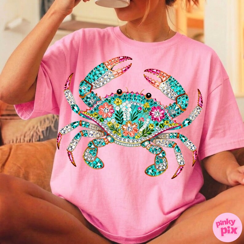 May include: Pink t-shirt featuring a detailed crab design. The crab is adorned with turquoise, pink, and clear crystal-like embellishments, and floral accents. The crab's claws are a mix of pink and orange. The shirt has a relaxed fit.