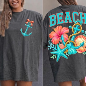 May include: Dark grey t-shirt with a turquoise anchor and orange bow on the front. The back features the word "BEACH" in turquoise letters, along with a floral and nautical design including an anchor, starfish, and seashells.