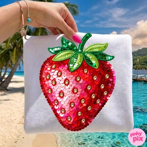 K&ouml;nnte beinhalten: Wei&szlig;es quadratisches Kissen mit einem gro&szlig;en, paillettenbesetzten Erdbeermotiv. Die Erdbeere ist rot mit rosa Schattierungen, gr&uuml;nen Bl&auml;ttern und goldfarbenen Edelsteinen. Das Bild ist vor einem tropischen Strandhintergrund.