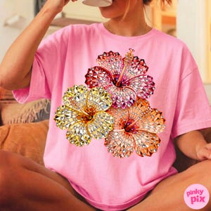 May include: Pink t-shirt featuring a vibrant floral design. The graphic showcases three hibiscus flowers in shades of yellow, orange, and red, with a sparkling, jeweled effect. The shirt is a casual, oversized style.