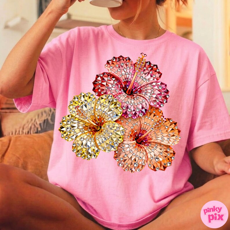 May include: Pink t-shirt featuring a vibrant floral design. The graphic showcases three hibiscus flowers in shades of yellow, orange, and red, with a sparkling, jeweled effect. The shirt is a casual, oversized style.