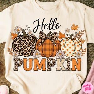 Op de afbeelding: Cr&egrave;mekleurig T-shirt met een herfstthema. Het ontwerp toont drie pompoenen met verschillende patronen, omringd door herfstbladeren en madeliefjes. De woorden "Hello PUMPKIN" zijn geschreven in een mix van lettertypen.