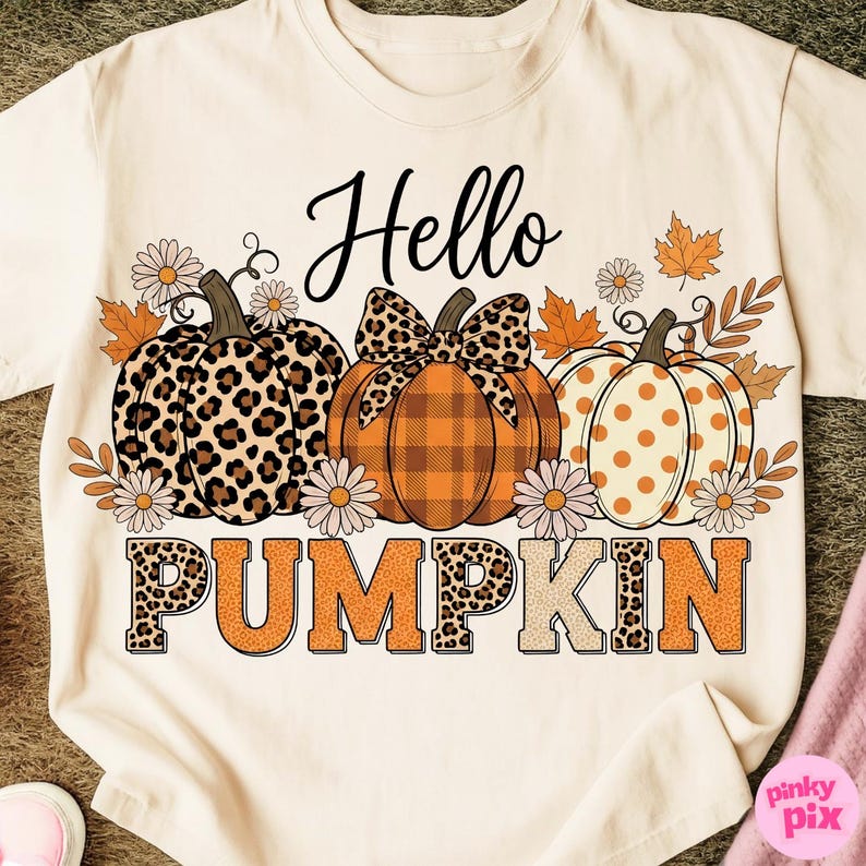 Op de afbeelding: Cr&egrave;mekleurig T-shirt met een herfstthema. Het ontwerp toont drie pompoenen met verschillende patronen, omringd door herfstbladeren en madeliefjes. De woorden "Hello PUMPKIN" zijn geschreven in een mix van lettertypen.
