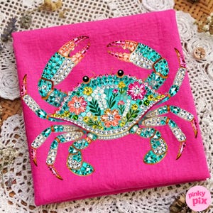 May include: A hot pink fabric features a detailed crab design. The crab is adorned with turquoise, orange, and clear crystal-like embellishments. Floral patterns and sparkling accents decorate the crab's body and claws, creating a vibrant and eye-catching design.