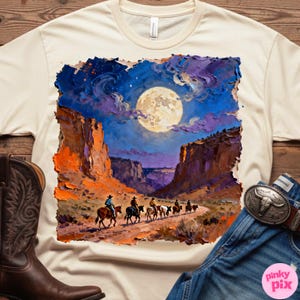 May include: Cream-colored t-shirt featuring a vibrant, impressionistic painting of cowboys riding horses through a canyon under a full moon. The artwork is framed by a rugged, orange and purple landscape. A brown cowboy boot and blue jeans are visible.