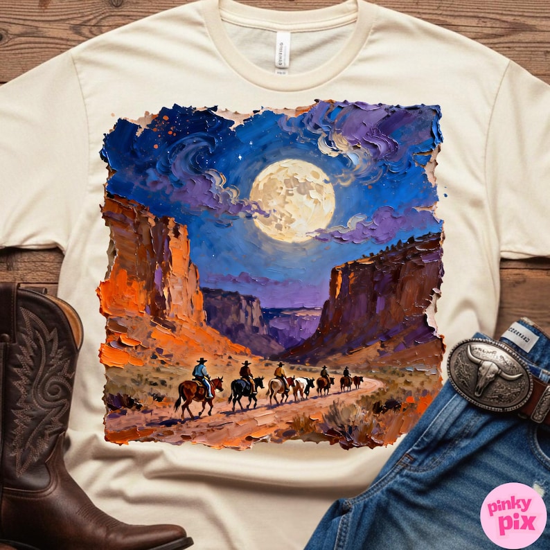May include: Cream-colored t-shirt featuring a vibrant, impressionistic painting of cowboys riding horses through a canyon under a full moon. The artwork is framed by a rugged, orange and purple landscape. A brown cowboy boot and blue jeans are visible.