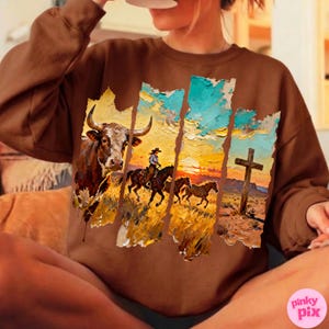 May include: Brown sweatshirt featuring a Western-themed graphic. The design includes a bull, a cowboy on horseback, and a cross against a sunset backdrop. The artwork has a textured, painted appearance.
