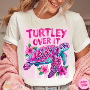 May include: White t-shirt featuring a colorful graphic of a sea turtle with the text "TURTLEY OVER IT". The turtle is pink, blue, and purple, surrounded by pink hibiscus flowers. The design is set against a pink striped background.