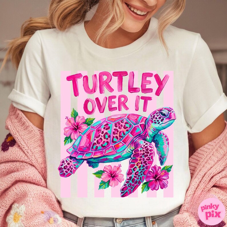 May include: White t-shirt featuring a colorful graphic of a sea turtle with the text "TURTLEY OVER IT". The turtle is pink, blue, and purple, surrounded by pink hibiscus flowers. The design is set against a pink striped background.