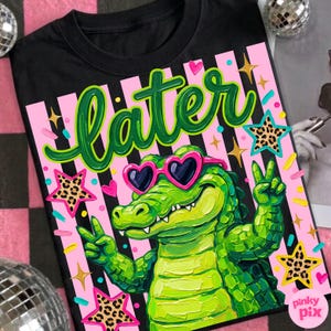 May include: Black t-shirt with a colorful graphic. The design features a green alligator wearing heart-shaped sunglasses, giving a peace sign, and the word "later" in green script. Pink and black stripes and star accents complete the design.