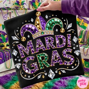May include: Black pillow with the words "MARDI GRAS" in purple and green jewel-like letters. The pillow features a jeweled crown and diamond accents. The design is held by a person with a purple sleeve. The background includes Mardi Gras beads and decorations.