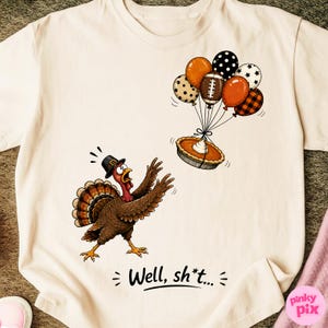 Pode incluir: Uma t-shirt cor de creme com um peru de desenho animado com um chapéu preto, tentando alcançar uma tarte de abóbora que flutua com um grupo de balões. Os balões têm padrões de bolinhas, estrelas e xadrez. O texto diz: Well, sh*t...