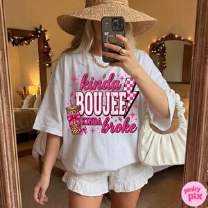 May include: A white short-sleeve t-shirt featuring a pink and black graphic design with the text "kinda BOUJEE kinda broke," a leopard print cup with a pink bow, a checkered lightning bolt, and small pink star accents throughout the print.