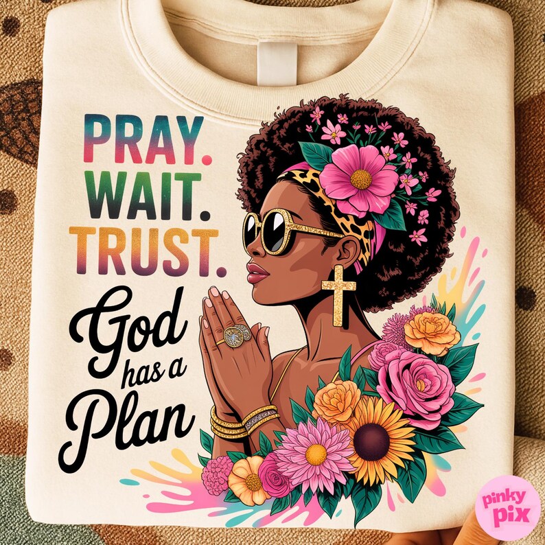 May include: Cream-colored sweatshirt featuring the text "PRAY. WAIT. TRUST. God has a Plan" in colorful lettering. The design includes an illustration of a woman with sunglasses, a cross earring, and floral accents.