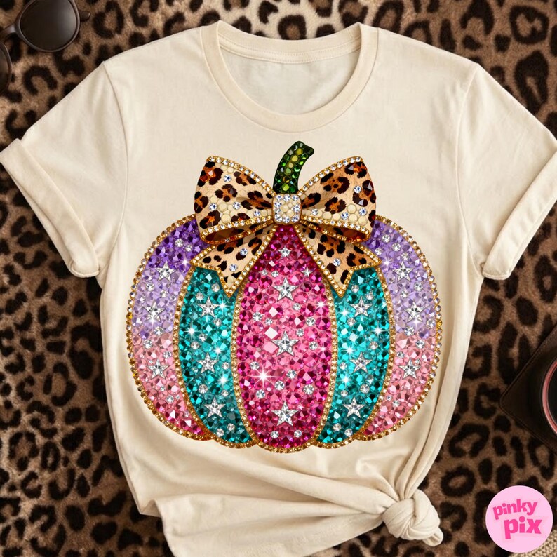 Op de afbeelding: Een cr&egrave;mekleurig t-shirt met korte mouwen en een grote afbeelding van een pompoen versierd met roze, turquoise en paarse gefacetteerde edelstenen en witte sterren. Een strik met luipaardprint en goudkleurige randen zit op de steel.