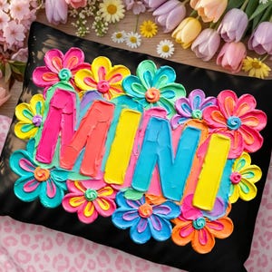 May include: Black decorative pillow with the word "MIMI" in colorful, textured paint. The letters are surrounded by bright, 3D flowers in shades of pink, yellow, blue, and orange. The pillow is on a pink and white patterned surface.